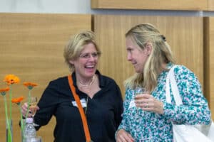 feldmann_robert_6320 | Quadriga Hochschule Berlin Zwei Frauen unterhalten sich lächelnd, eine hält ein Glas Sekt, die andere ein Glas Wasser, im Hintergrund orangefarbene...