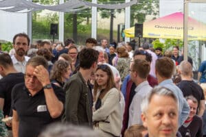 feldmann_robert_6382 | Quadriga Hochschule Berlin Menschenmenge bei einer Veranstaltung im Freien, mit Stehtischen und Sonnenschirmen im Hintergrund.