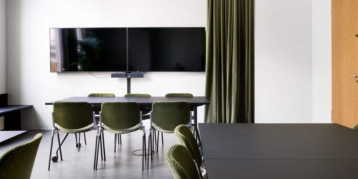 Moderne Besprechungsraum mit schwarzen Tischen, grünen Stühlen, einem großen Bildschirm und einer Pflanze in der Ecke.