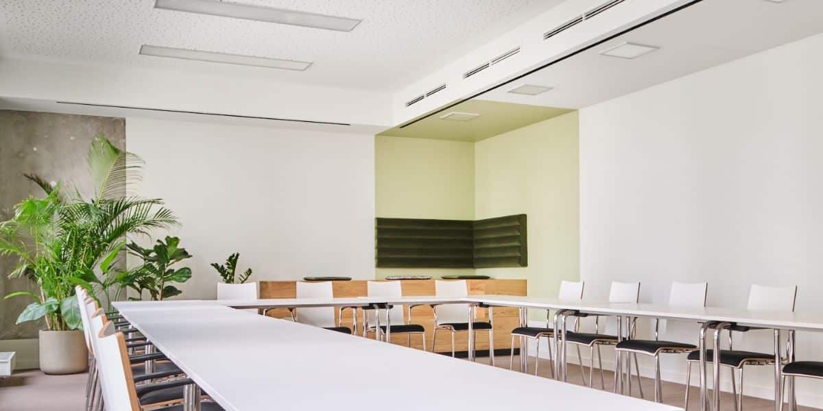 Heller Konferenzraum mit langem Tisch, weißen Stühlen und Pflanzen in den Ecken.