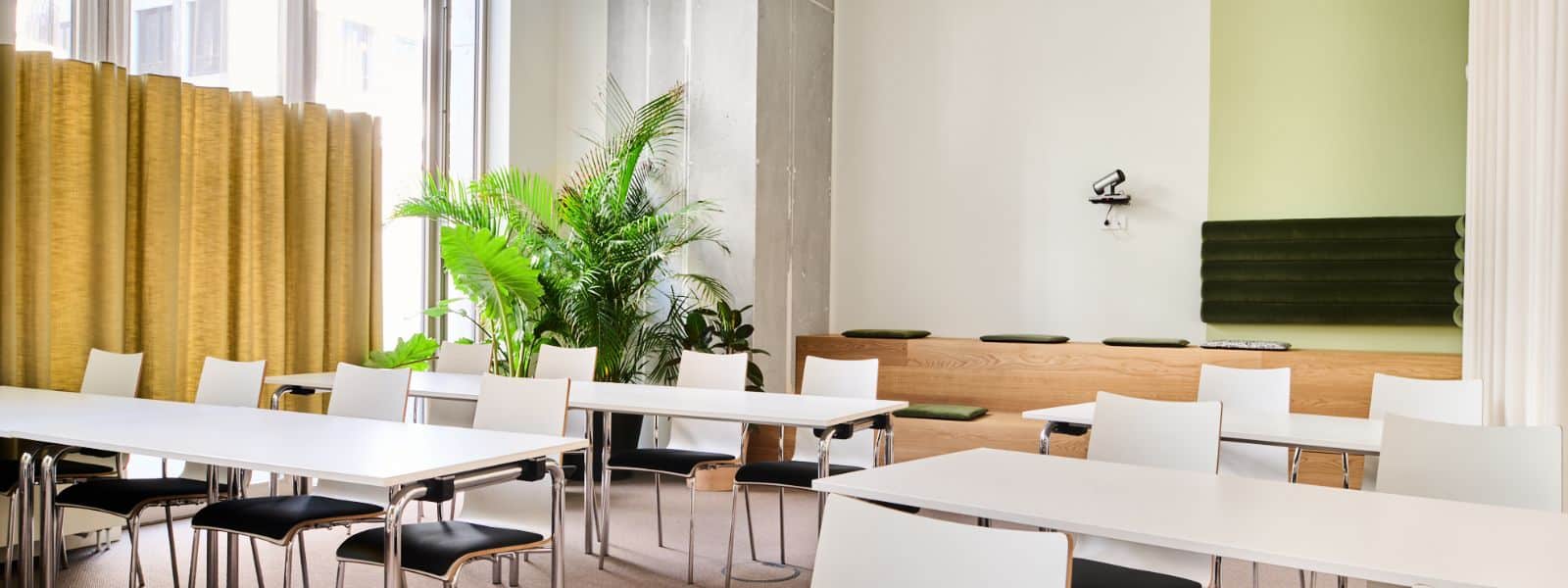 31 Heller Seminarraum mit weißen Tischen, schwarzen Stühlen, Pflanzen und einer grünen Wand.