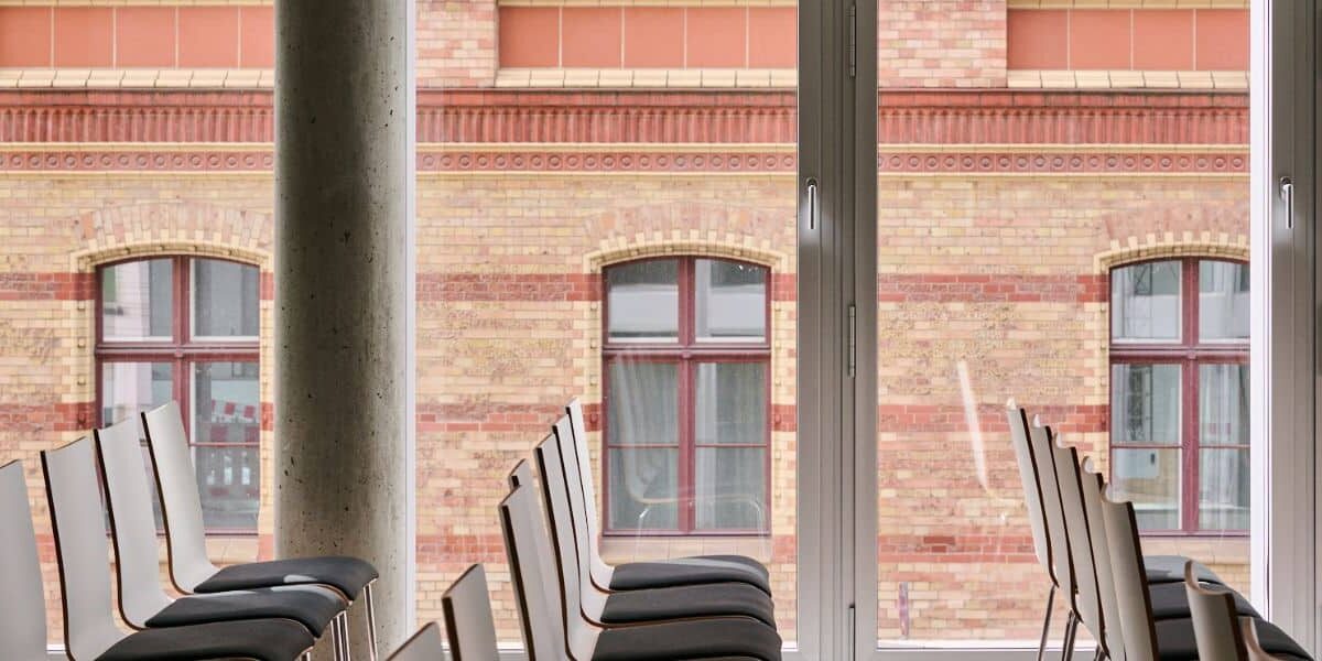 Leere Stühle stehen in einem modernen Raum mit großen Fenstern, die auf eine historische Backsteinfassade blicken.