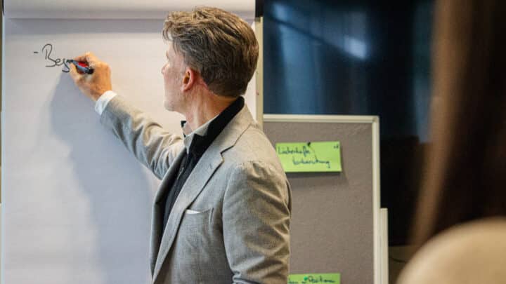 feldmann_robert_1536 Mann in grauem Anzug schreibt mit einem Marker auf ein Flipchart in einem Besprechungsraum.