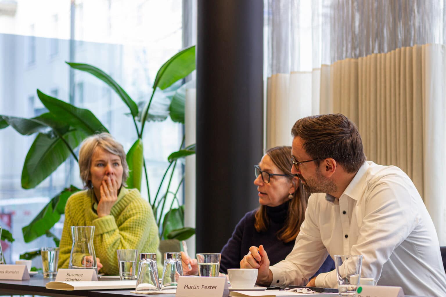 Drei Personen sitzen an einem Tisch, umgeben von Pflanzen, und diskutieren in einem hellen Raum.