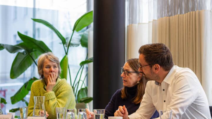 feldmann_robert_1584 Drei Personen sitzen an einem Tisch, umgeben von Pflanzen, und diskutieren in einem hellen Raum.